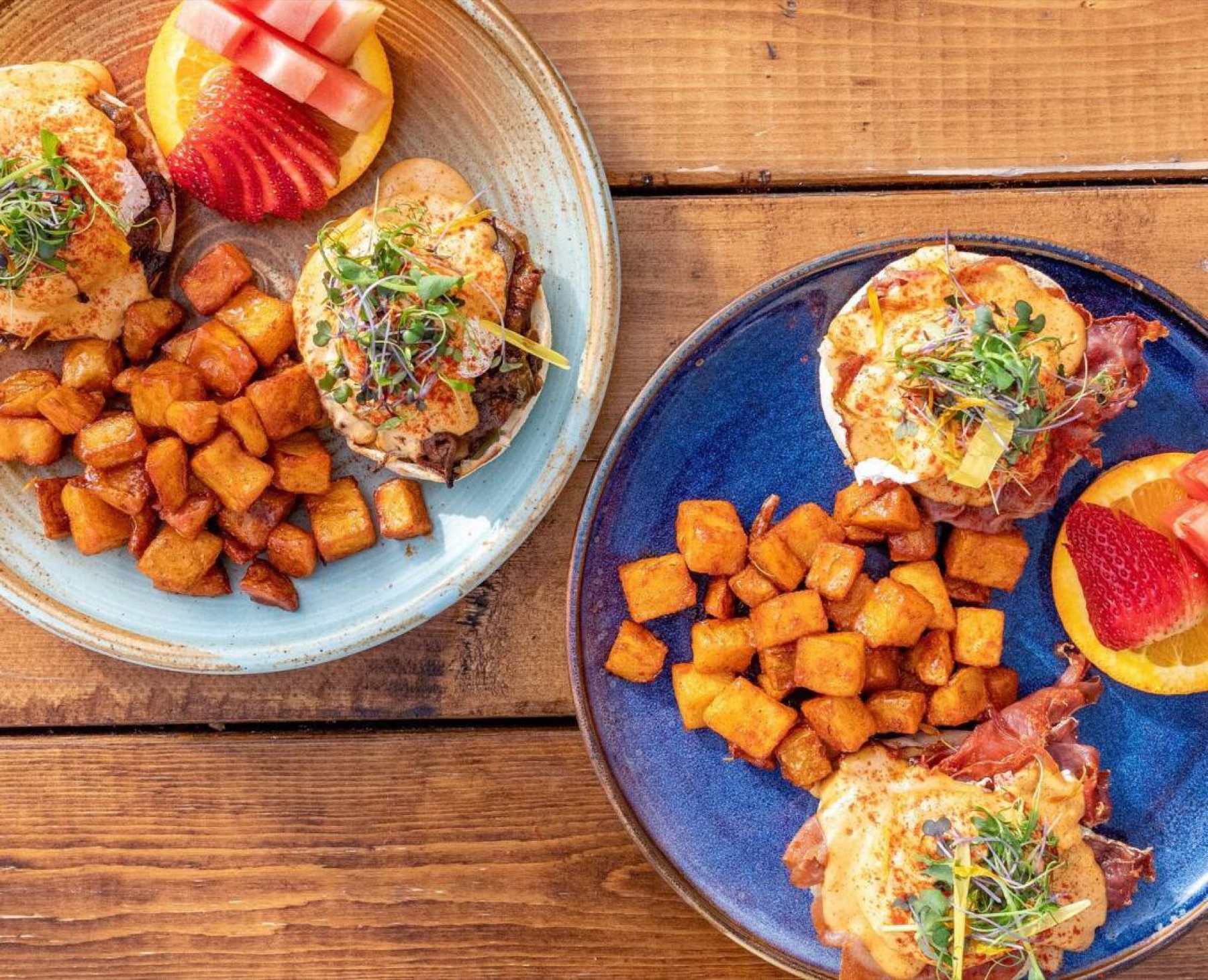 Two plates of Tiramisu Bistro eggs benedict with maple-spiced potatoes, fruit, and microgreens