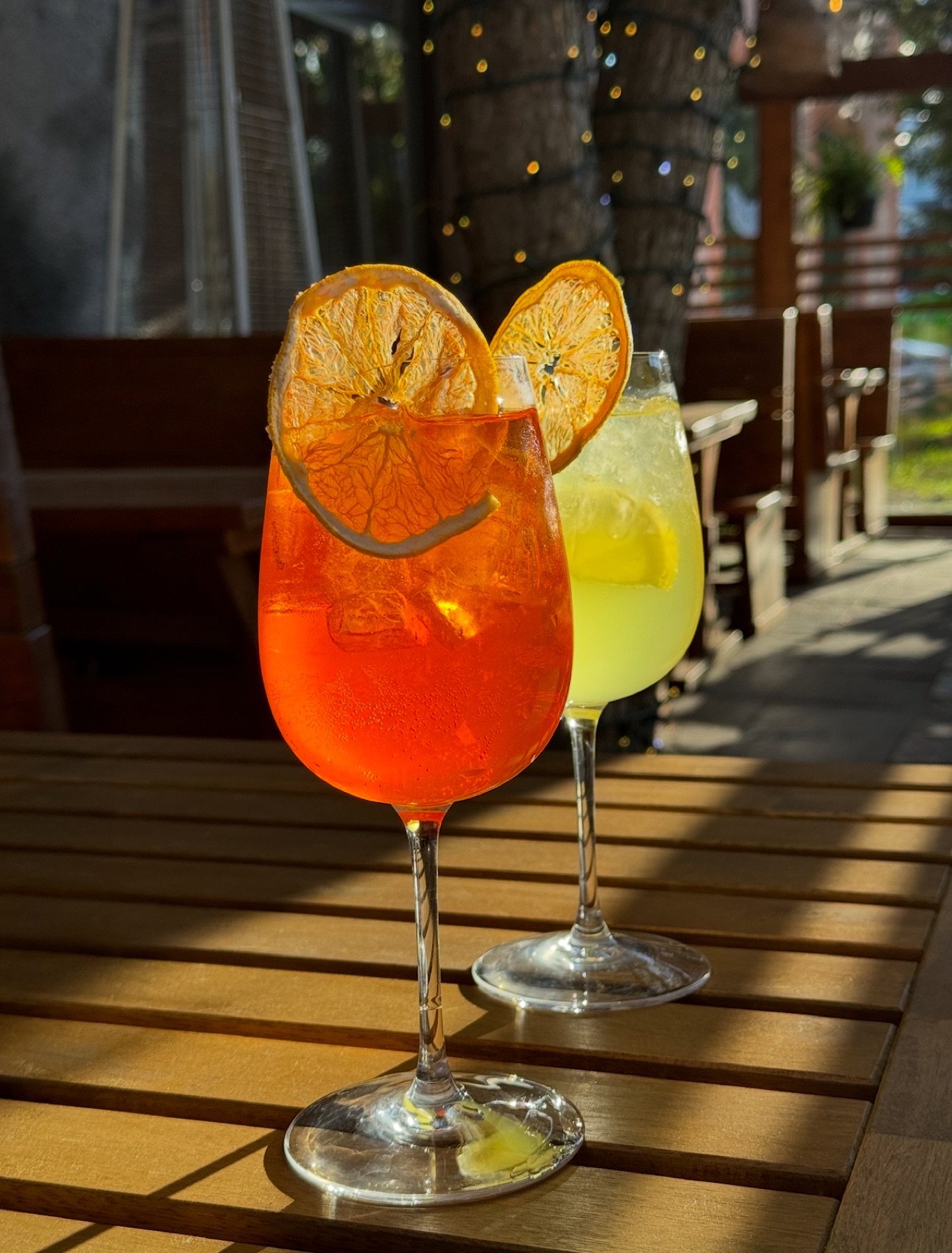 Aperol Spritz and a lemon cocktail with dehydrated citrus garnishes on a sunlit wooden patio table